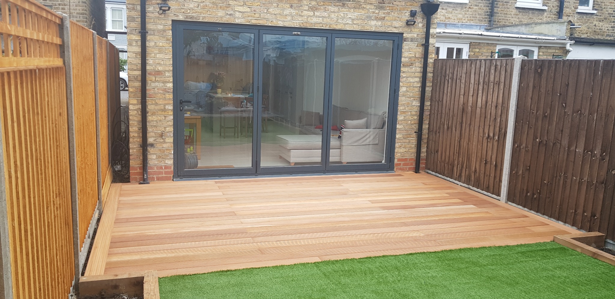 Nowoczesny taras z jasnego drewna przylegający do domu z dużymi przesuwnymi drzwiami, otoczony płotem i pasem sztucznej trawy.