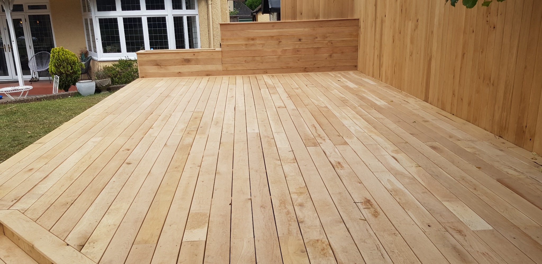 Nowoczesny taras drewniany z jasnego drewna z niskim ogrodzeniem z desek, widok na dom z białymi oknami.