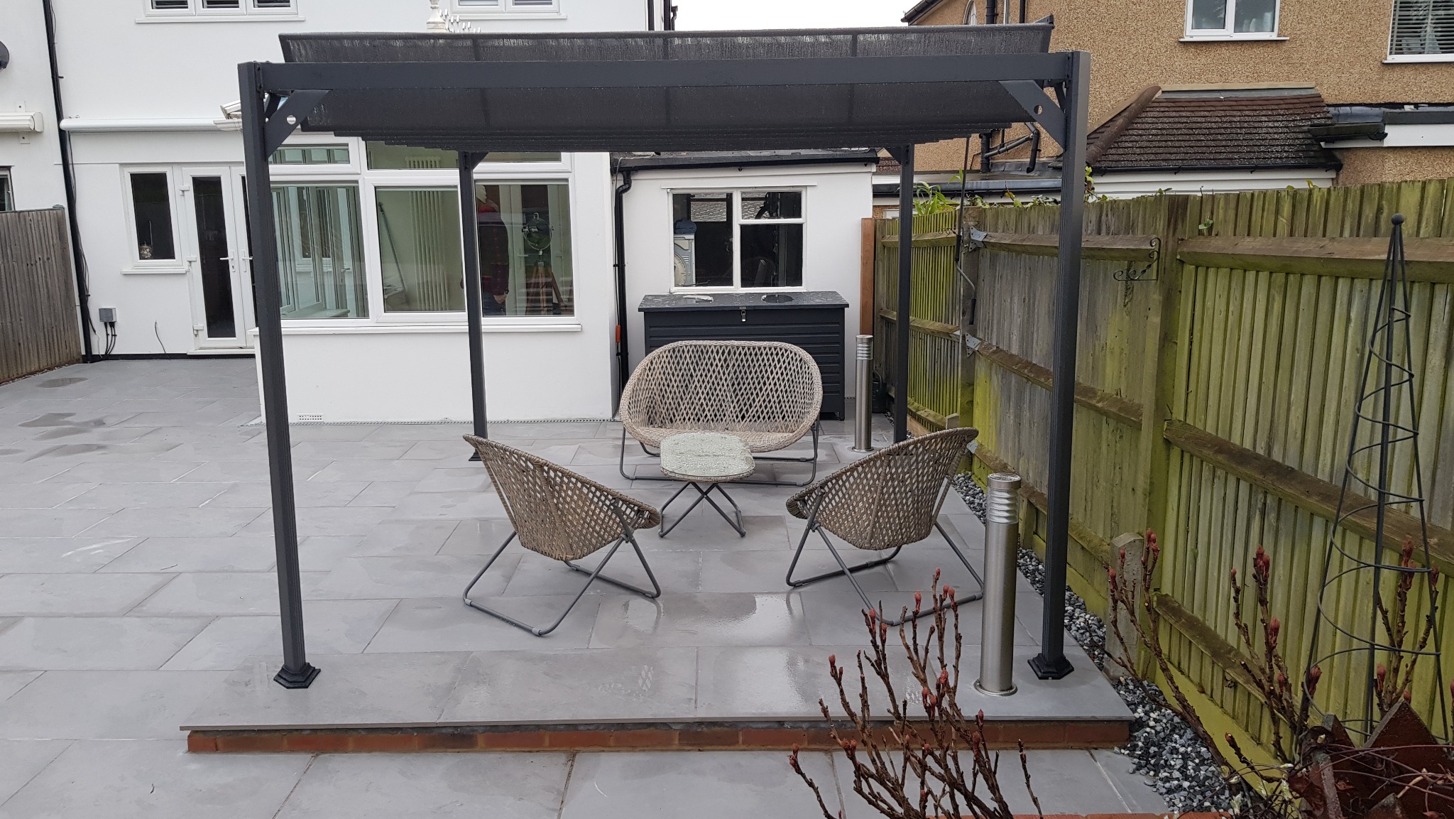 Nowoczesna metalowa pergola z zadaszeniem, pod nią rattanowe meble ogrodowe: sofa i dwa krzesła, na szarym tarasie z kamiennych płyt, w tle biały dom z dużymi oknami i drewniane ogrodzenie.