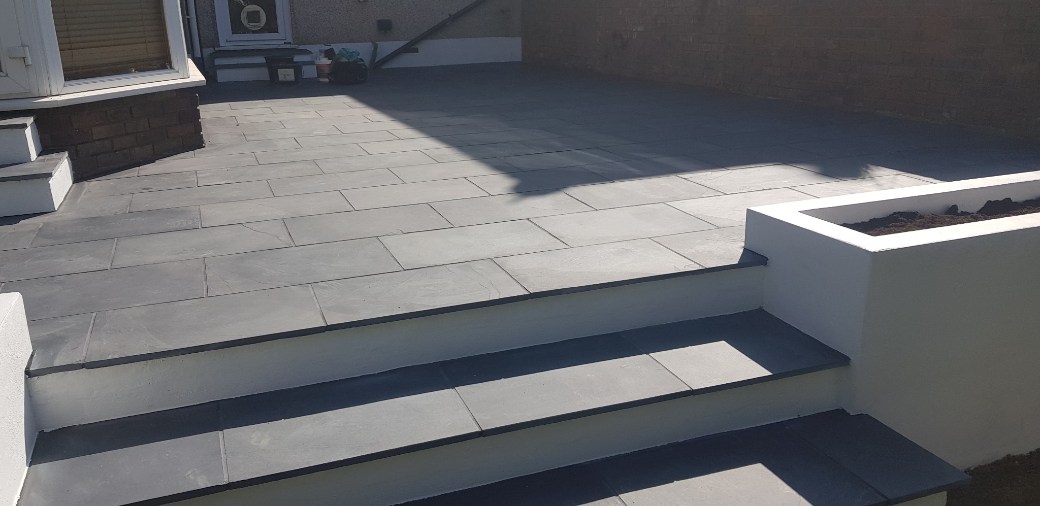 Nowoczesny taras z szarej kamiennej kostki, schody z kamienia prowadzące do domu, biała donica z ziemią w tle, ceglany mur.