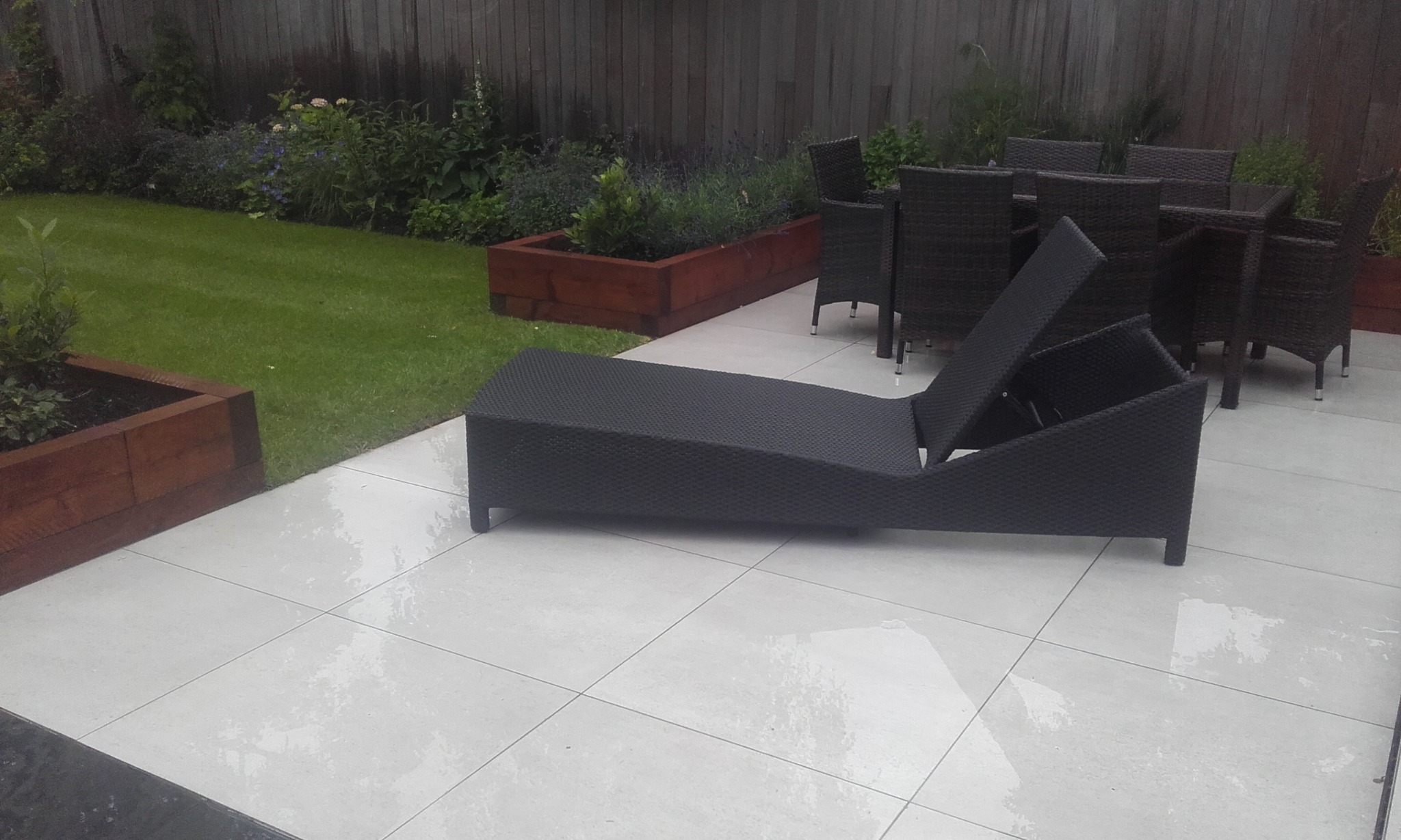 Biały taras z dużych płyt, z czarnym leżakiem i zestawem mebli ogrodowych z rattanu, na tle zielonego trawnika i drewnianego ogrodzenia.