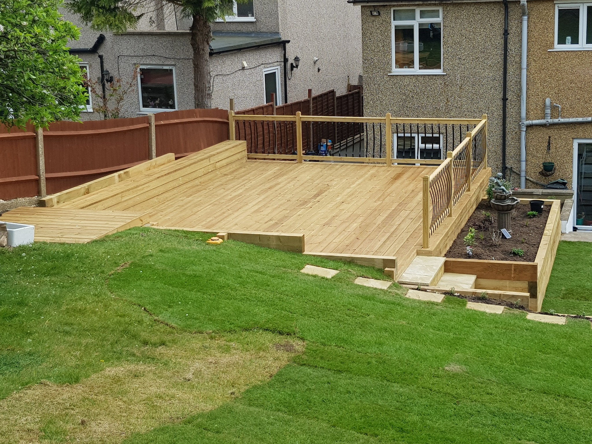 Nowoczesny taras z drewna z balustradą i schodami, wkomponowany w zielony trawnik z elementami ogrodu z fontanną.