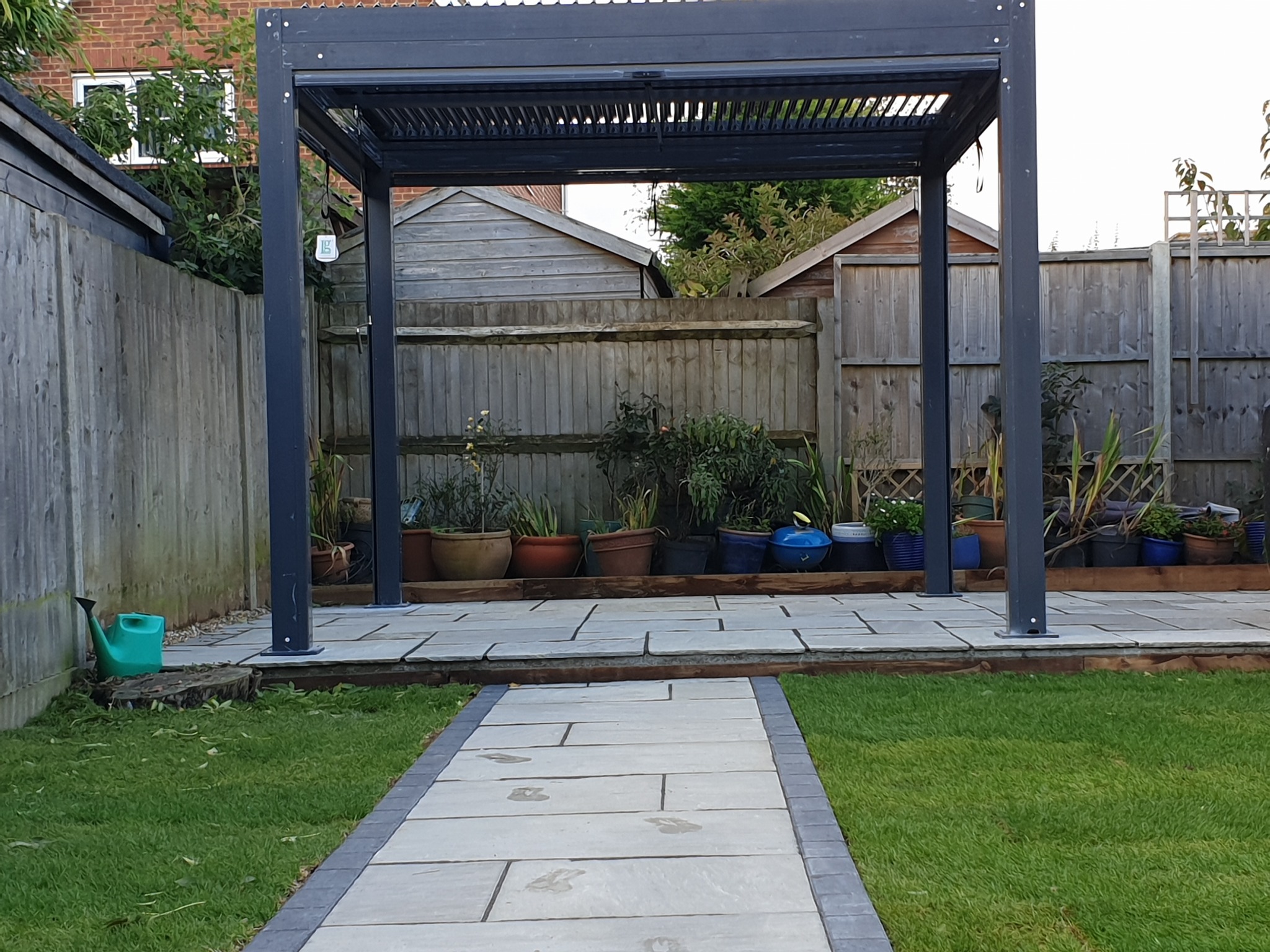 Nowoczesna, metalowa pergola w ogrodzie z kamienną ścieżką, doniczkami z roślinami i drewnianym ogrodzeniem w tle.