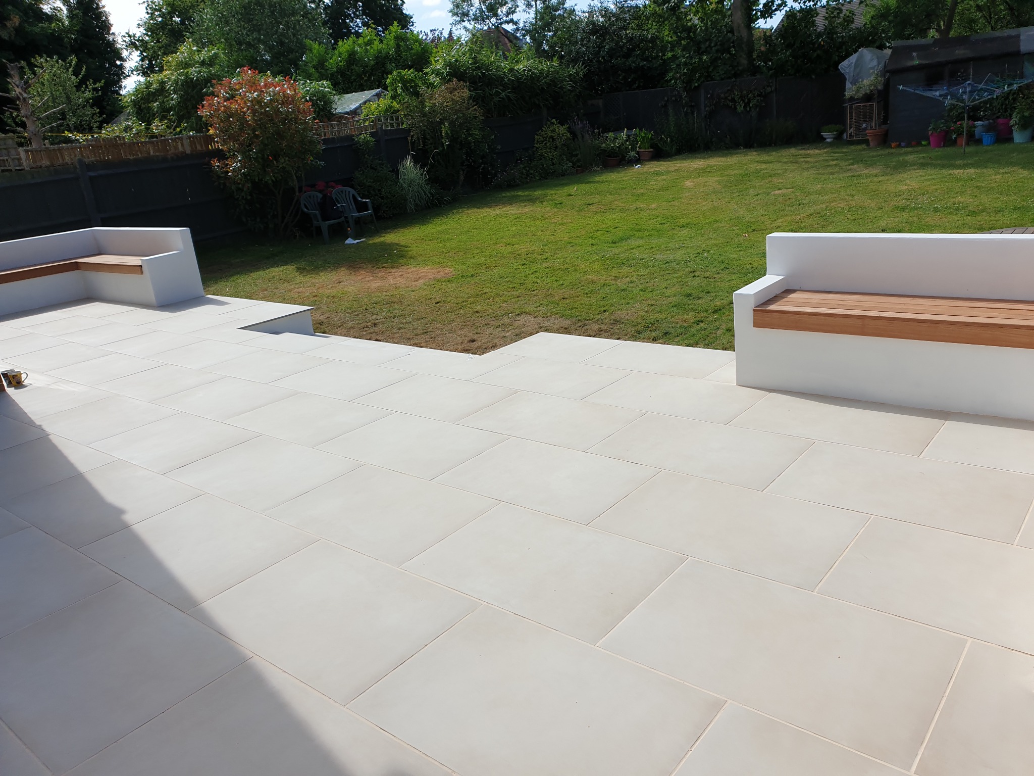 Jasny taras z dużych płyt z wbudowanymi siedziskami z drewnianymi blatami, widok na trawnik i ogrodzenie.