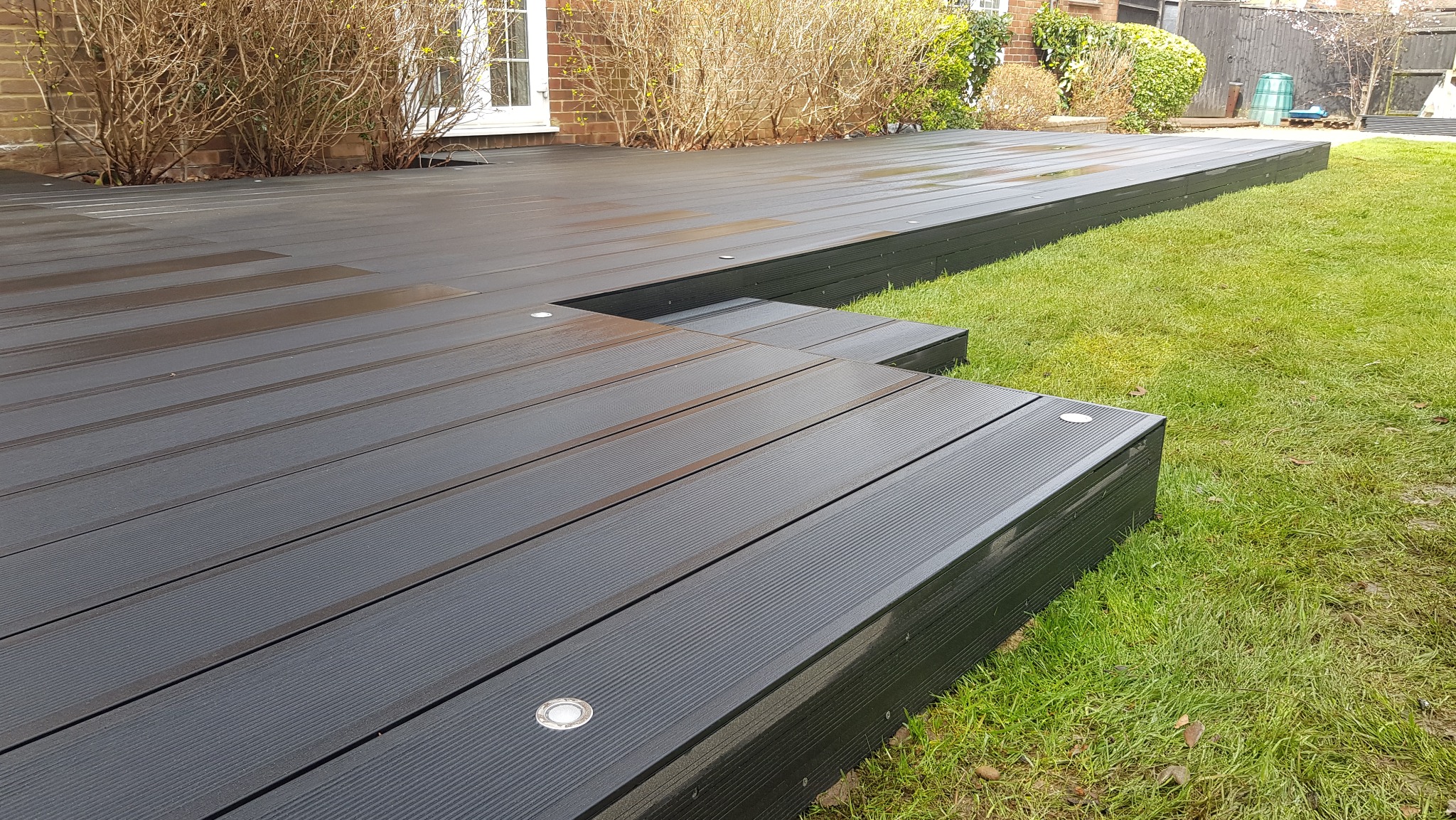 Czarny taras kompozytowy z wbudowanym oświetleniem punktowym, przylegający do trawnika i ceglanego budynku, częściowo mokry po deszczu.