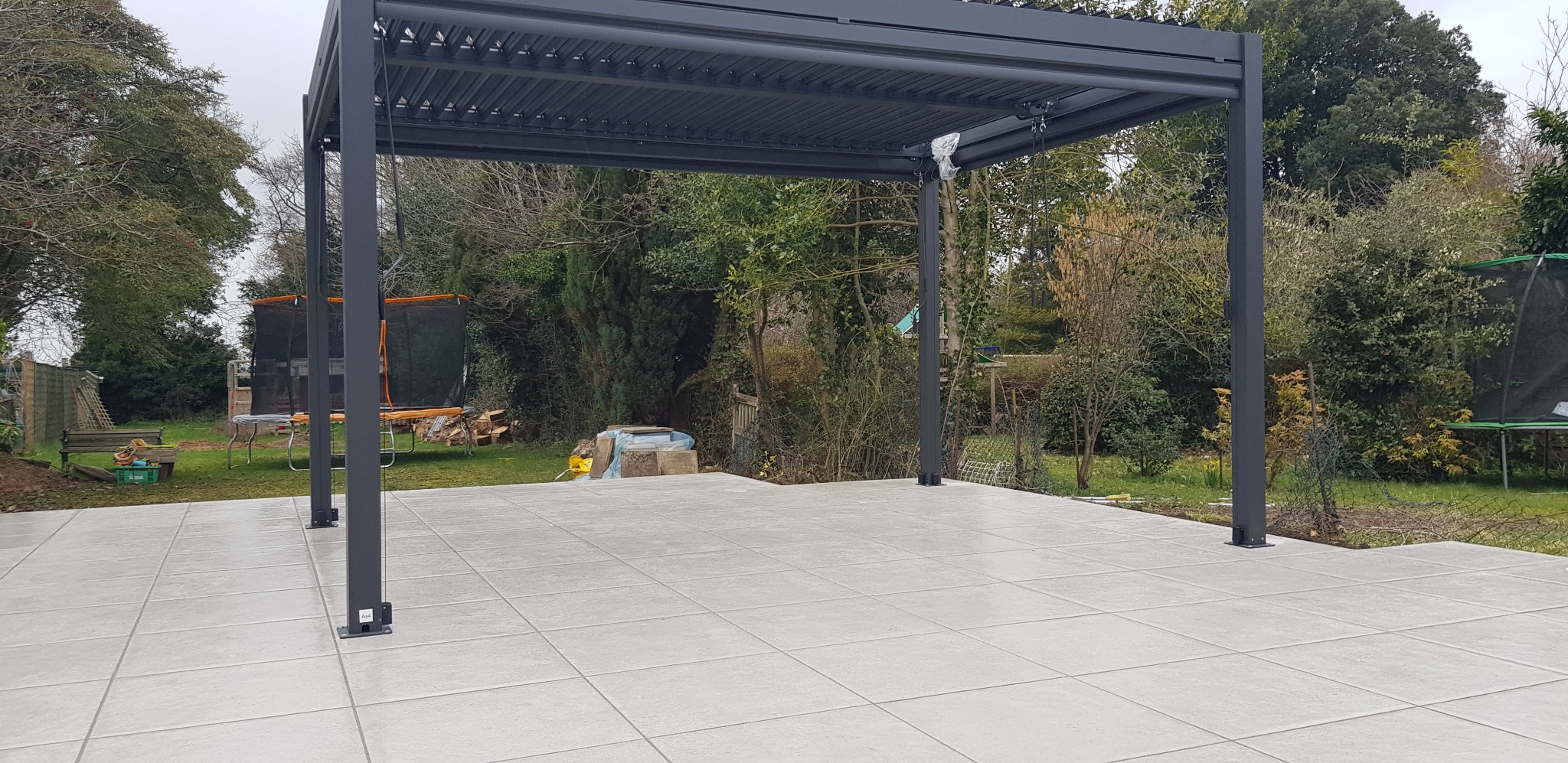 Nowoczesna pergola metalowa z regulowanym zadaszeniem, ustawiona na jasnym tarasie z płytek, w tle zieleń ogrodu.