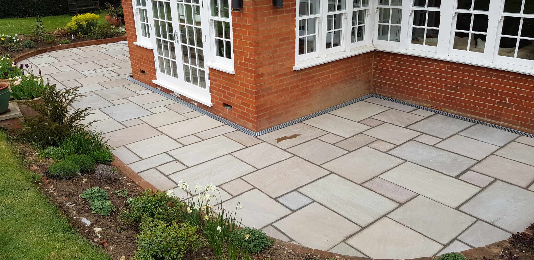 Jasny taras z kamiennych płyt ułożonych w nieregularny wzór, przylegający do ceglanego budynku z białymi oknami, obrzeża z cegły i rabaty z roślinami.