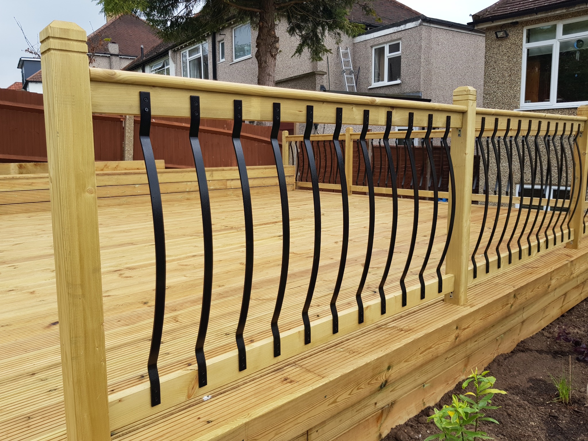 Nowy taras z jasnego drewna z czarnymi, metalowymi balustradami o falistym kształcie, widok z poziomu gruntu na tle domów jednorodzinnych.