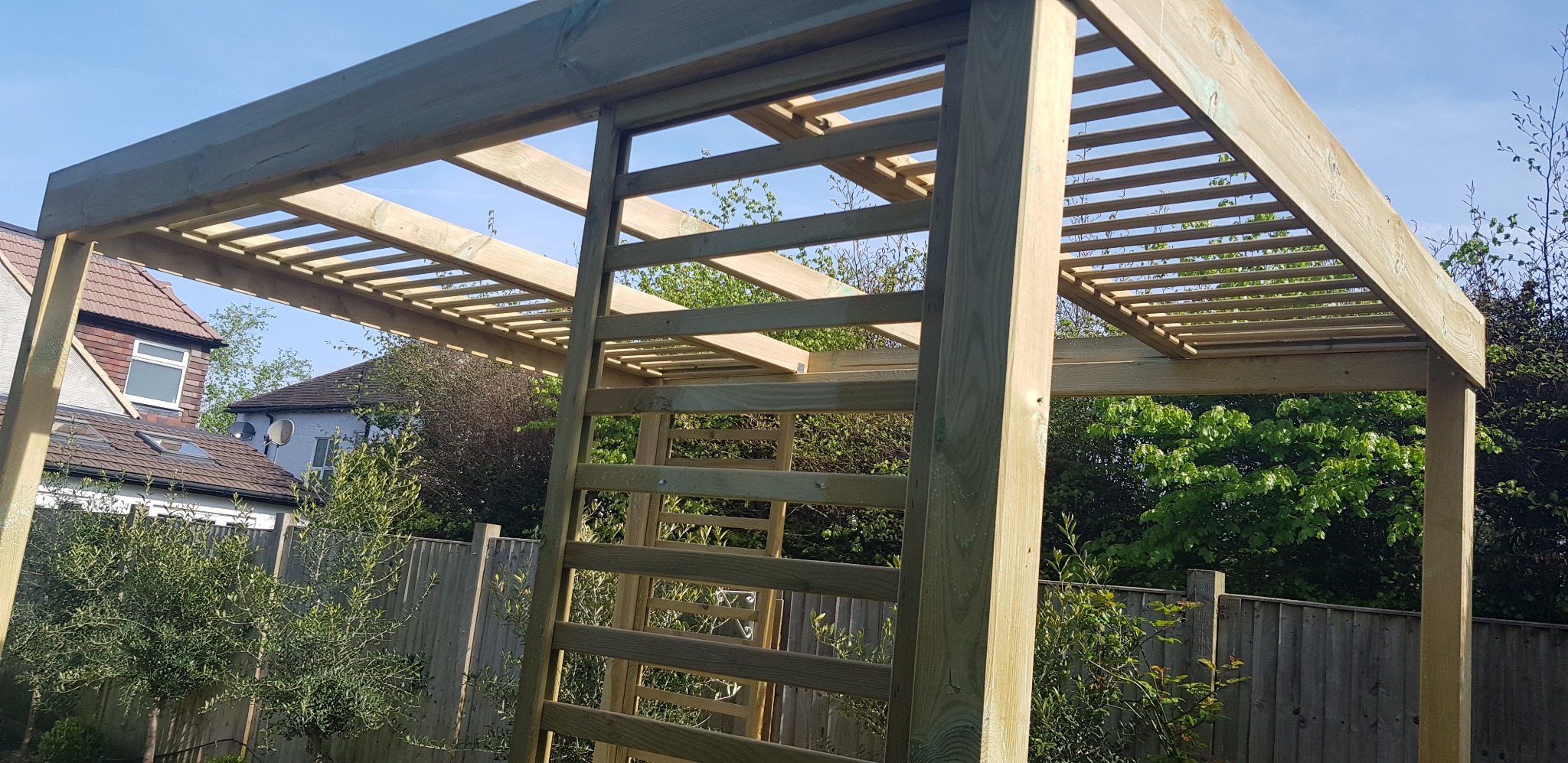 Surowa, drewniana pergola w trakcie montażu, widoczne elementy konstrukcyjne i jasne drewno na tle zieleni ogrodowej i zabudowań.