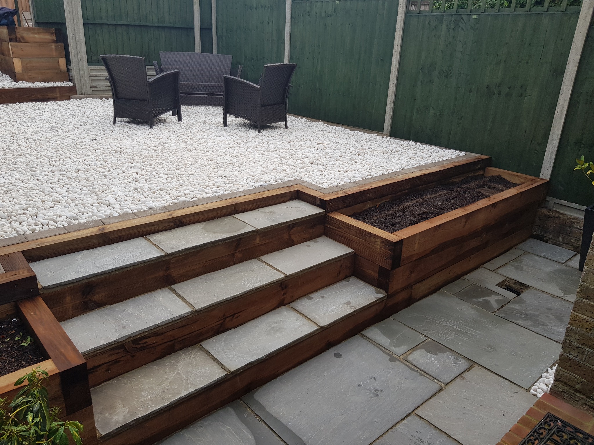 Ogród z tarasem wyłożonym szarymi płytami, drewnianymi schodami i strefą relaksu z białymi kamykami oraz meblami ogrodowymi z technorattanu.