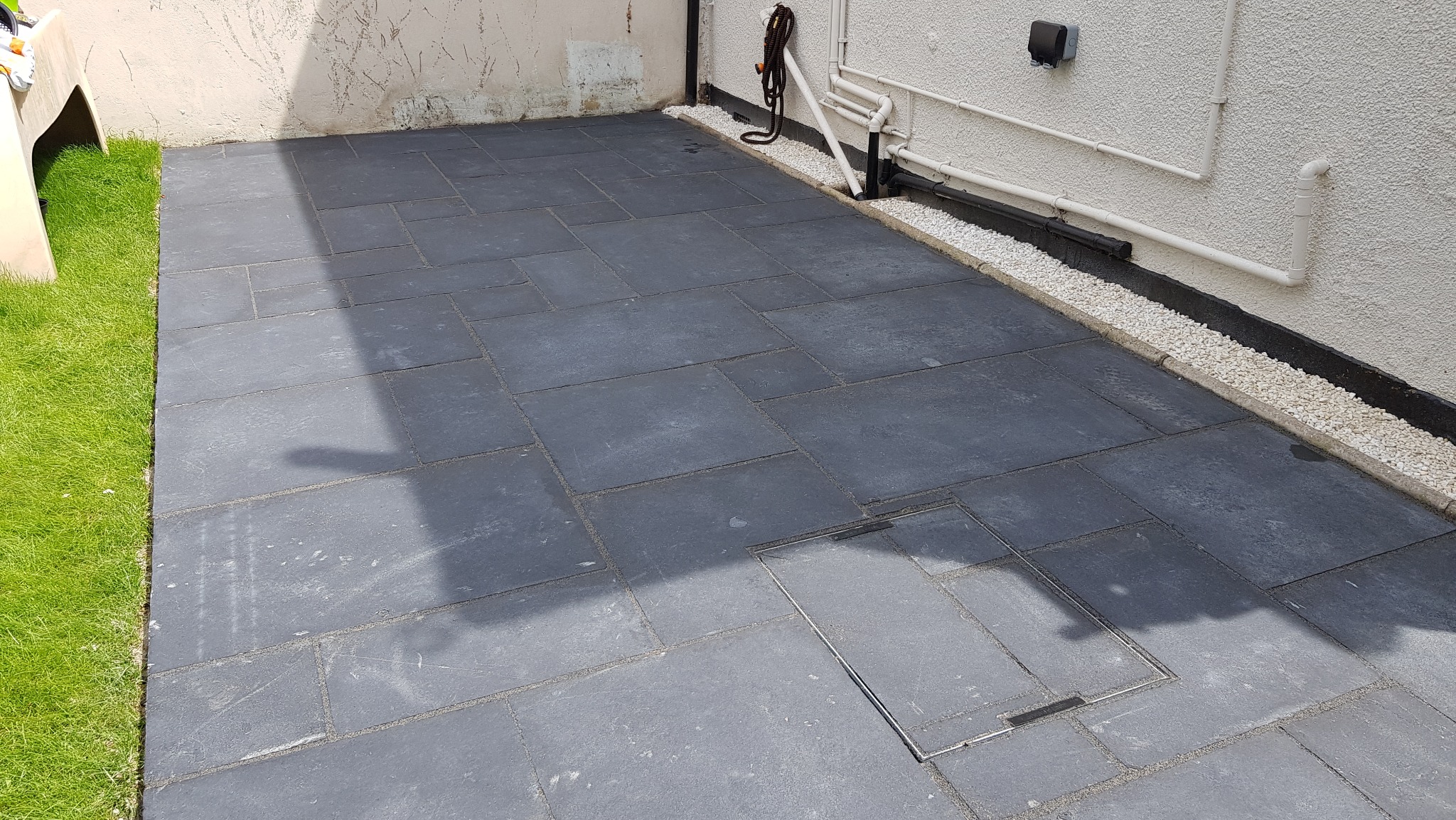 Nowoczesny taras wyłożony ciemnoszarymi płytami kamiennymi o zróżnicowanych wymiarach, z widoczną studzienką rewizyjną i obrzeżem z białego grysu przy ścianie budynku.