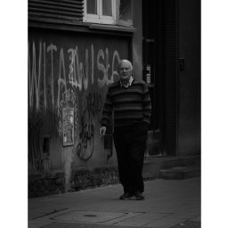 IgorBaścikFoto - Czarno-białe zdjęcie starszego mężczyzny idącego chodnikiem przed budynkiem pokrytym graffiti, z ręką w kieszeni i papierosem w dłoni.