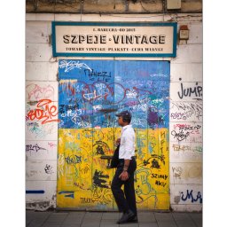 IgorBaścikFoto - Mężczyzna w słuchawkach przechodzi obok drzwi pomalowanych w barwy flagi Ukrainy, pokrytych graffiti, z szyldem 'Szpeje Vintage: Towary Vintage, Plakaty, Cuda Wianki'.