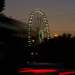 IgorBaścikFoto - Oświetlone koło diabelskie w nocy, z rozmazanymi śladami świateł samochodów na dole i zarysem autobusu w tle.