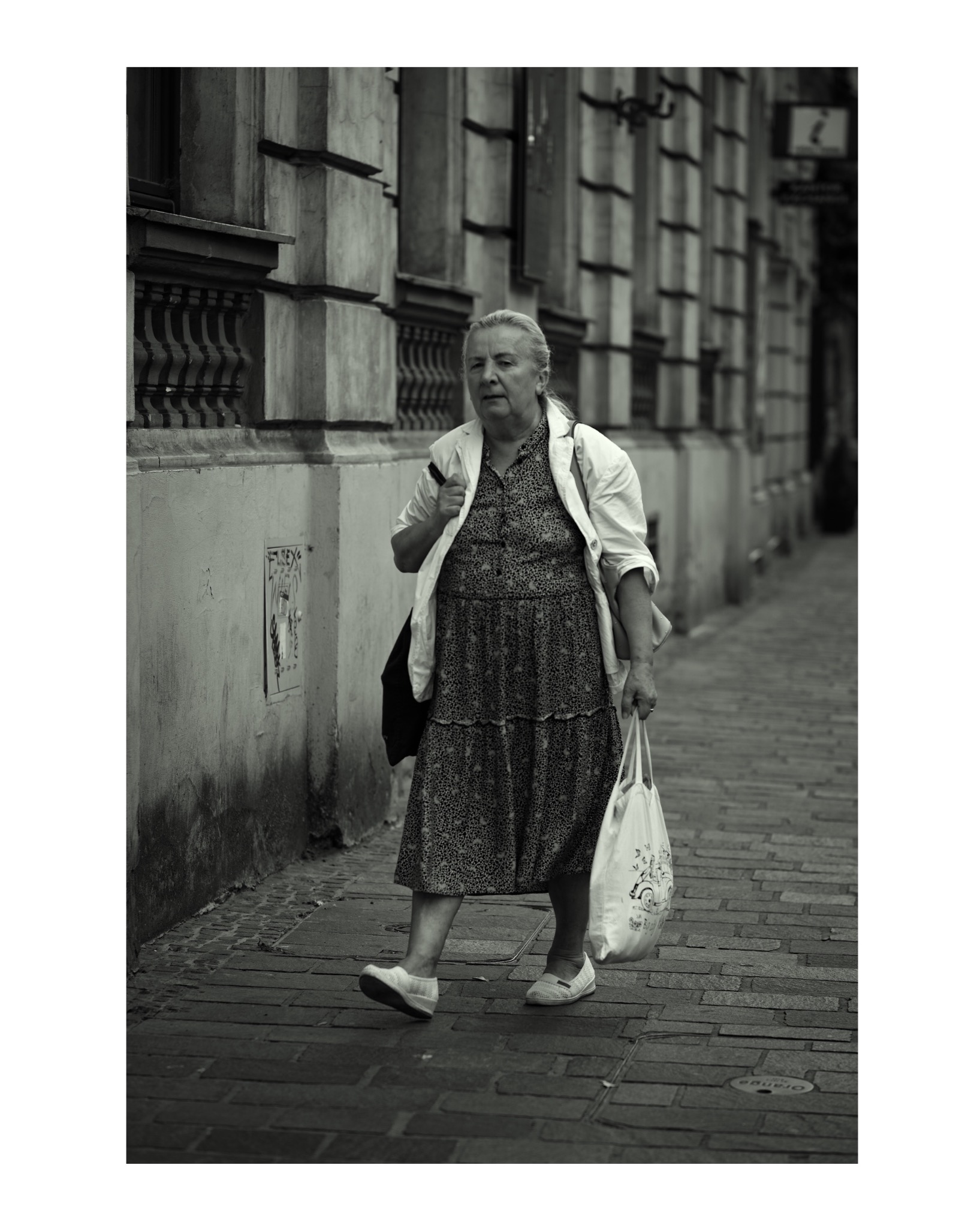 Czarno-białe zdjęcie kobiety idącej chodnikiem obok kamienicy, trzymającej torbę na zakupy.