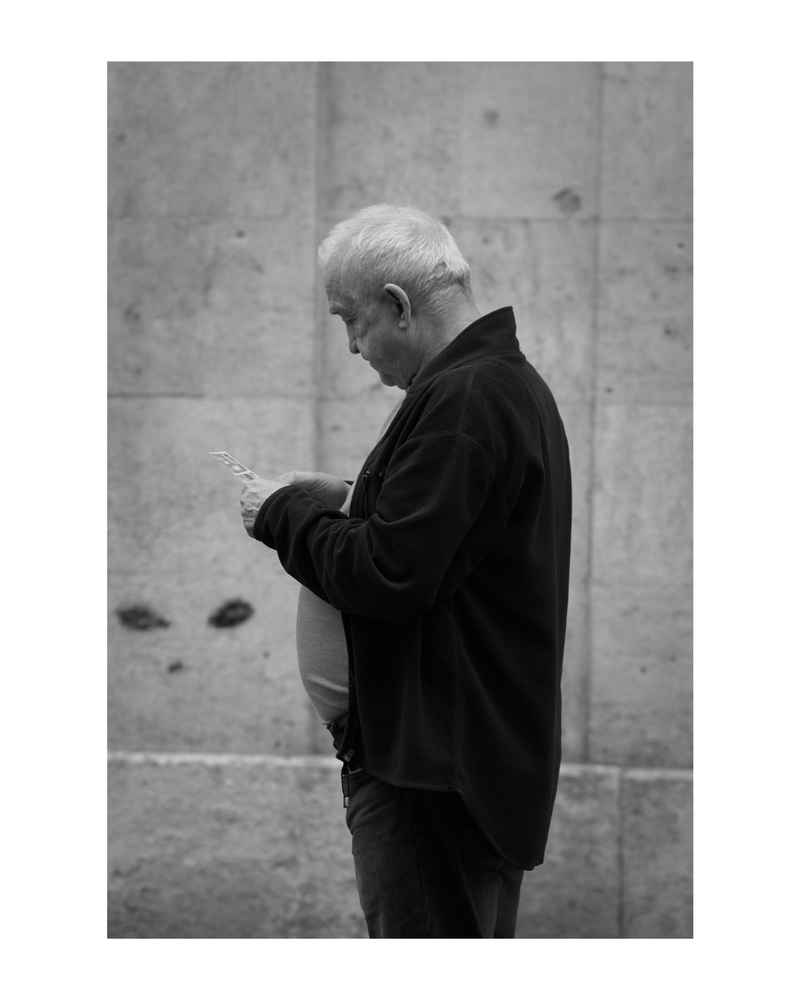 Czarno-białe zdjęcie starszego mężczyzny w ciemnej bluzie, czytającego ulotkę na tle betonowej ściany.