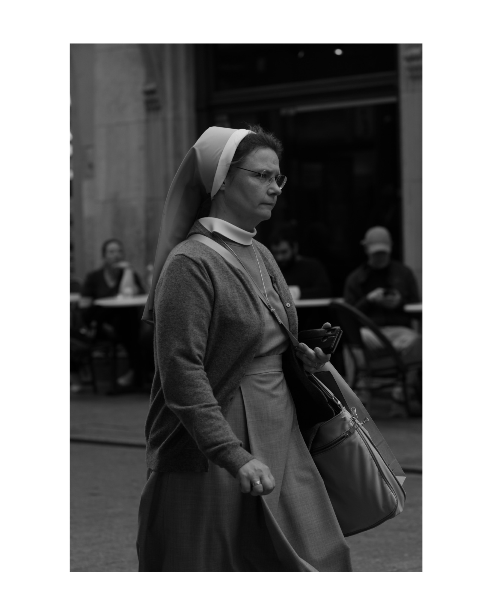 Czarno-białe zdjęcie zakonnicy w okularach, idącej ulicą z telefonem i torbą na ramieniu, w tle stoliki kawiarniane