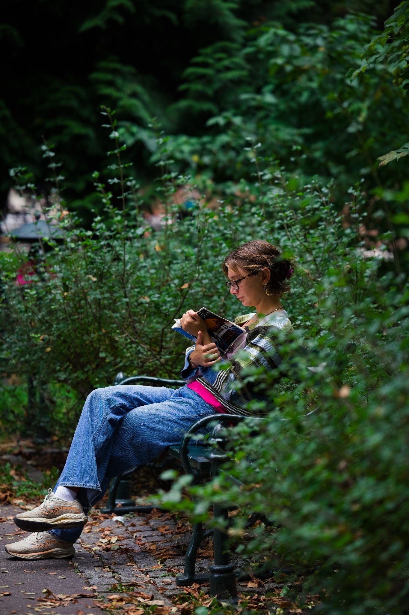 Kobieta w okularach siedzi na ławce w parku, czytając komiks z okładką w odcieniach niebieskiego i pomarańczowego, otoczona zielenią krzewów.