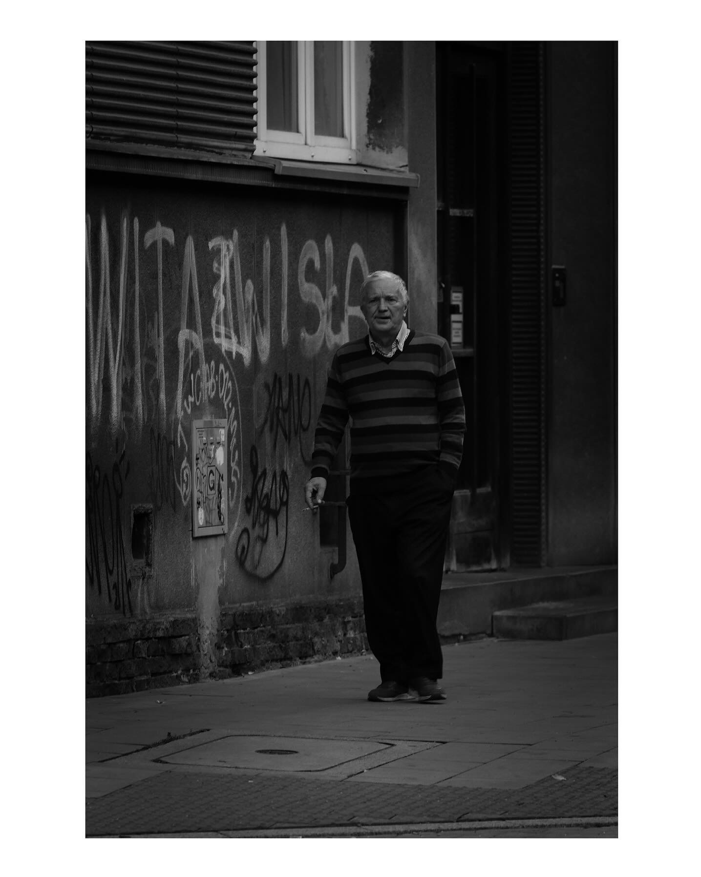 Czarno-białe zdjęcie starszego mężczyzny idącego chodnikiem przed budynkiem pokrytym graffiti, z ręką w kieszeni i papierosem w dłoni.