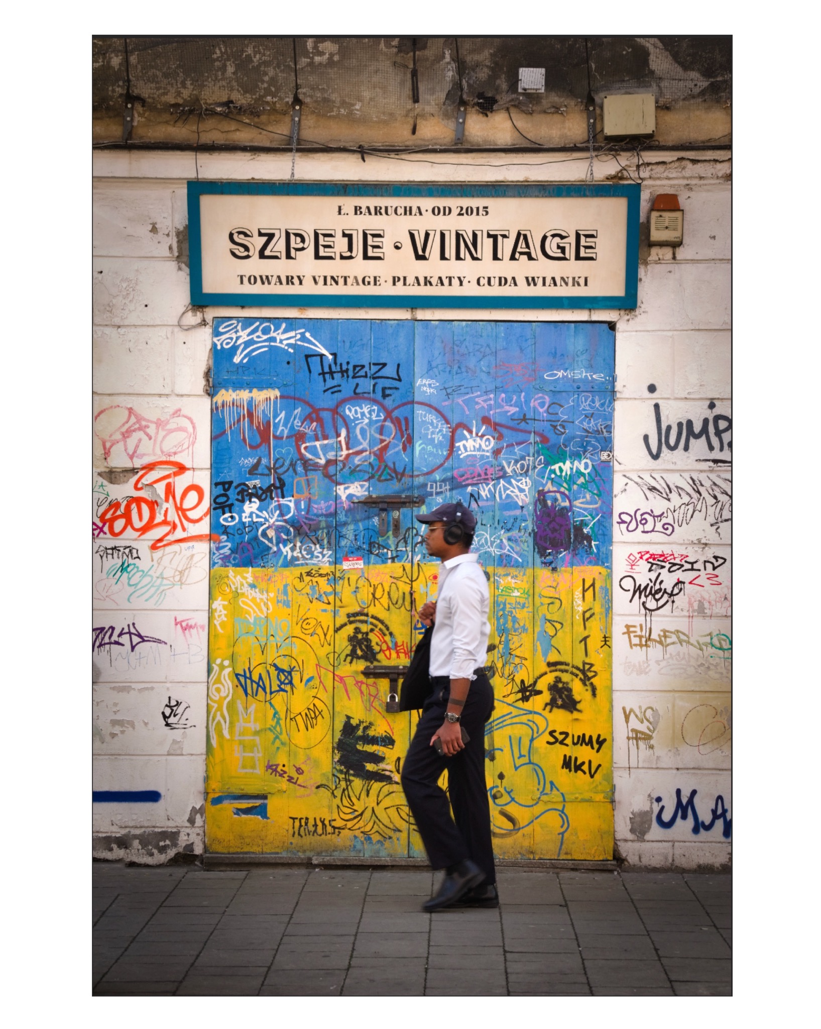 Mężczyzna w słuchawkach przechodzi obok drzwi pomalowanych w barwy flagi Ukrainy, pokrytych graffiti, z szyldem 'Szpeje Vintage: Towary Vintage, Plakaty, Cuda Wianki'.