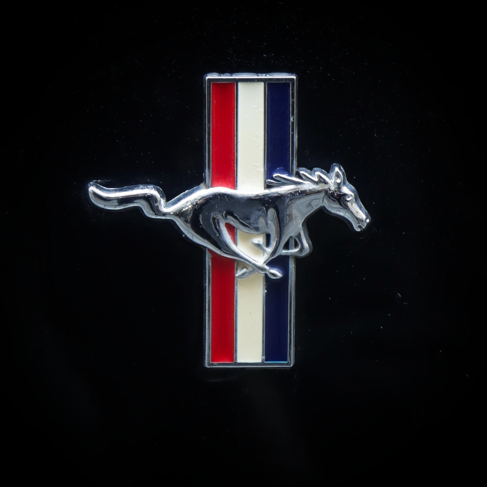 Chromowany emblemat galopującego konia na tle pionowych pasów w kolorach czerwonym, białym i niebieskim, umieszczony na czarnej powierzchni.