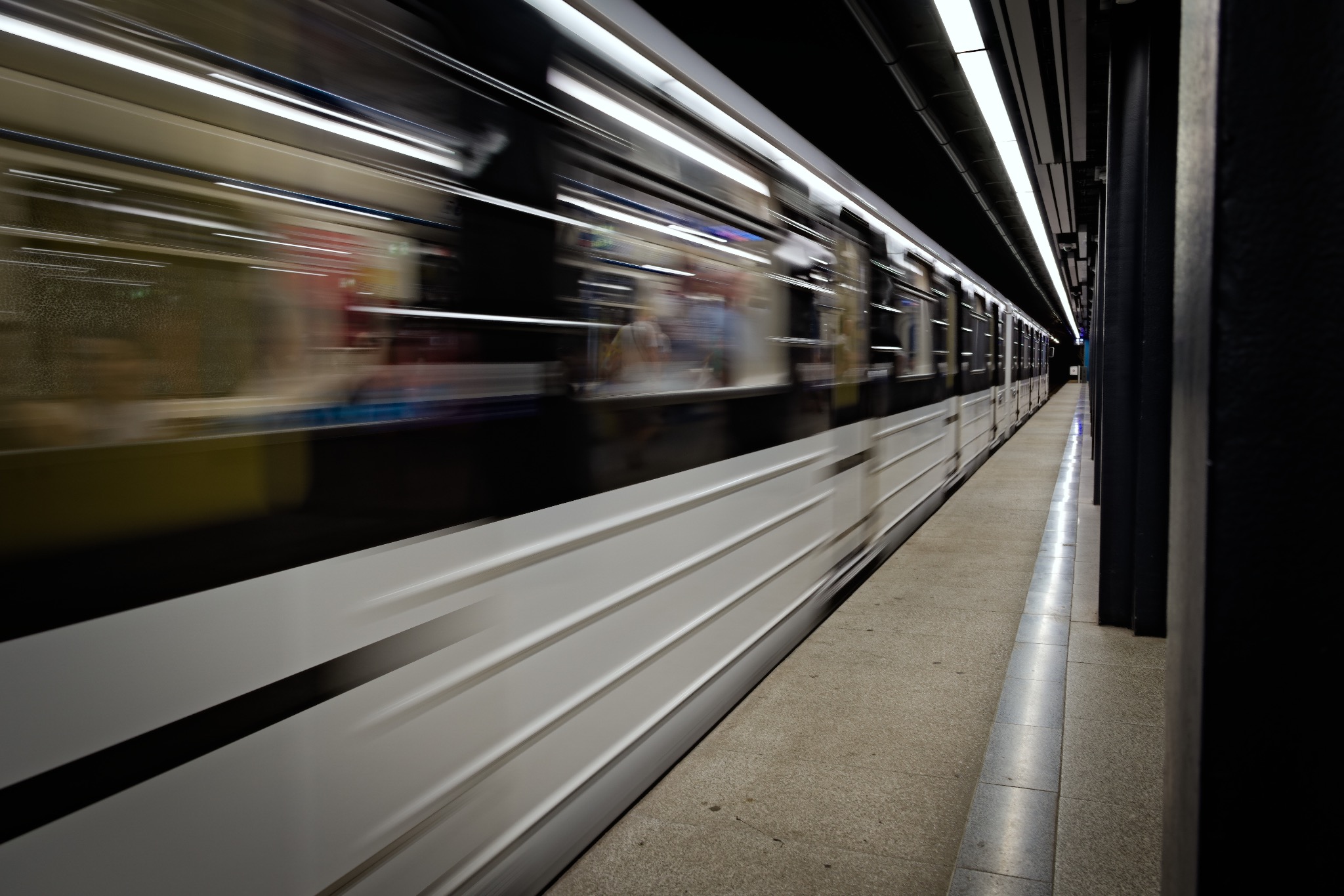 Rozmyte ujęcie pędzącego wagonu metra na stacji, z widocznymi światłami sufitowymi i fragmentem peronu