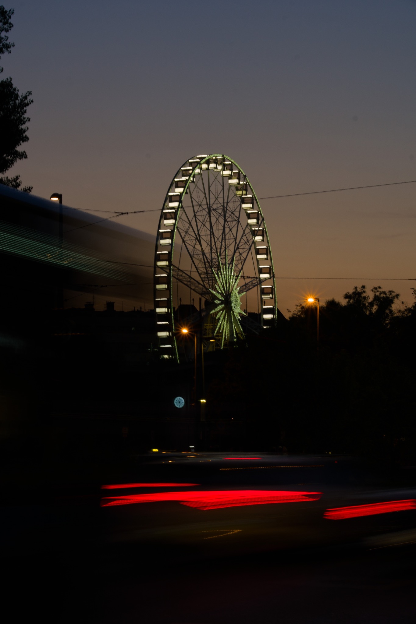 Oświetlone koło diabelskie w nocy, z rozmazanymi śladami świateł samochodów na dole i zarysem autobusu w tle.