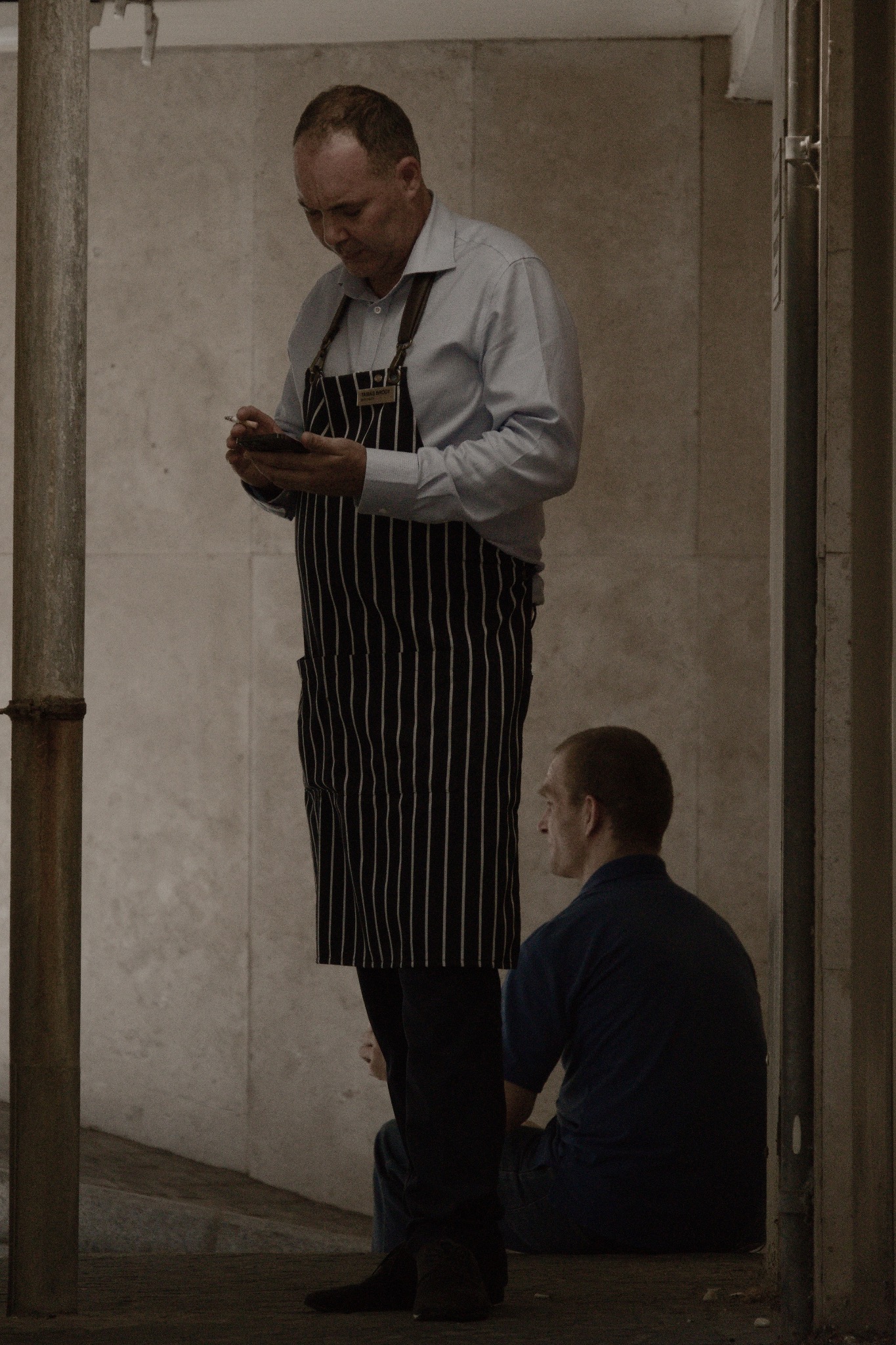 Kelner w pasiastej zapasce sprawdza zamówienie na smartfonie, w tle siedzący mężczyzna w niebieskiej koszuli.