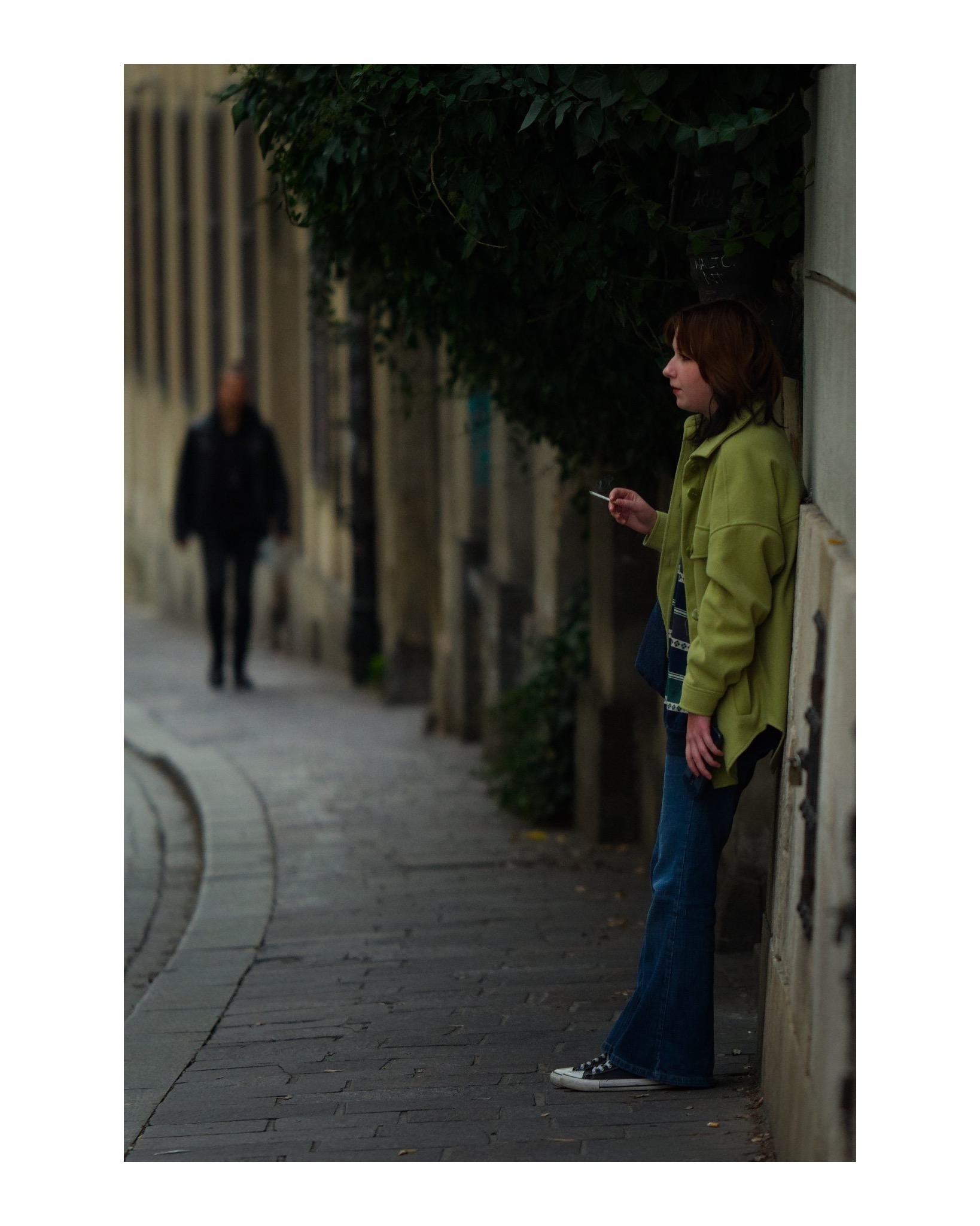 Kobieta w zielonej kurtce oparta o ścianę na ulicy, trzymająca papierosa, w tle zamazana postać przechodnia.