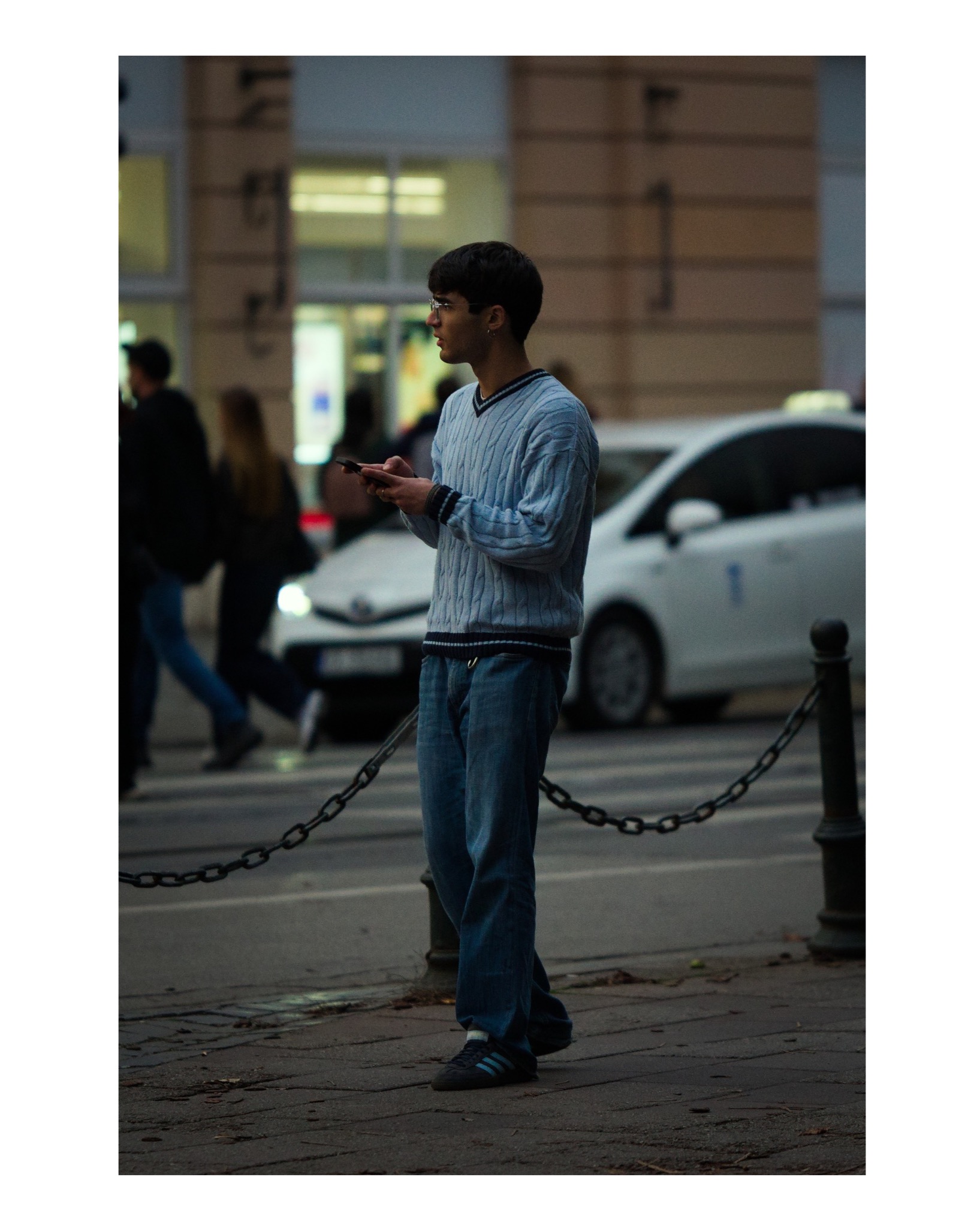 Młody mężczyzna w okularach, ubrany w błękitny sweter i dżinsy, stoi na chodniku i patrzy w dal, trzymając telefon komórkowy; w tle ruch uliczny i budynki miejskie.