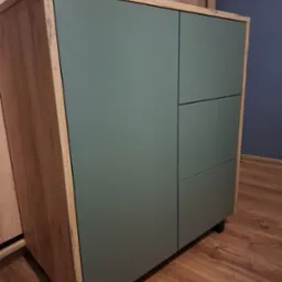 Niska szafka z dwiema pionowymi szafkami i trzema szufladami w kolorze zielonym, z obudową z drewna w stylu rustykalnym, na tle drewnianej podłogi i ściany w odcieniach niebieskiego.