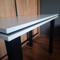 Patryk Ziemba - Biały stół z czarnymi nogami na tle szafy z drewnianymi frontami i szarej ściany, widoczne detale konstrukcyjne blatu.