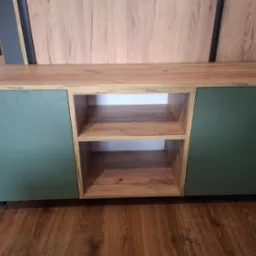 Nowoczesna szafka RTV z drewnianym blatem i zielonymi frontami, widoczne dwie otwarte półki i brązowa podłoga.