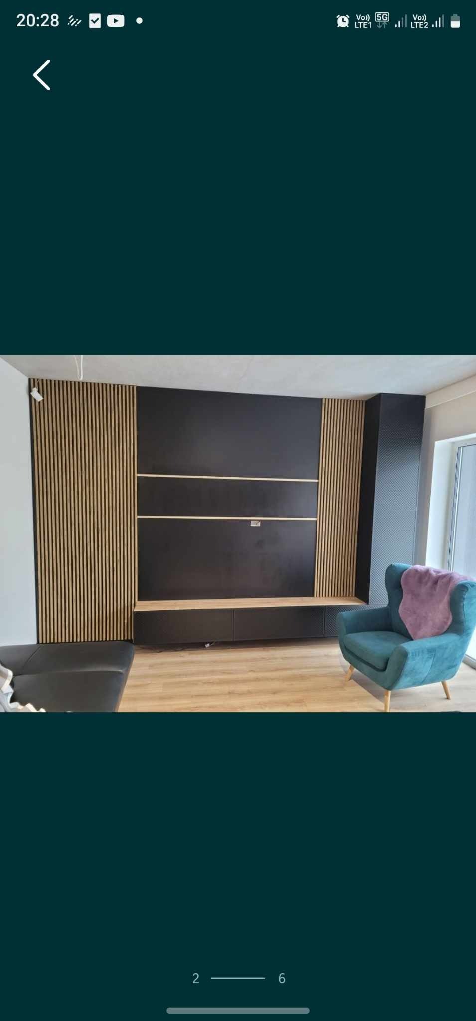 Czarna ściana telewizyjna z drewnianymi lamelami po bokach, pod nią szafka RTV, na podłodze jasny panel, w rogu pokoju turkusowy fotel z narzutą.