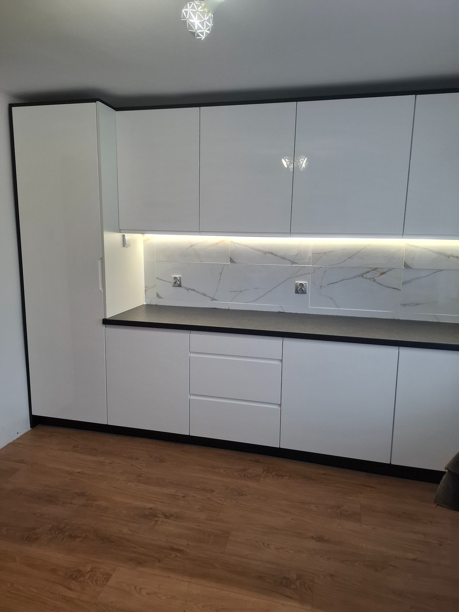 Nowoczesna, biała zabudowa kuchenna z czarnym blatem i marmurowym panelem nad blatem, oświetlonym listwą LED, widok na całą ścianę.