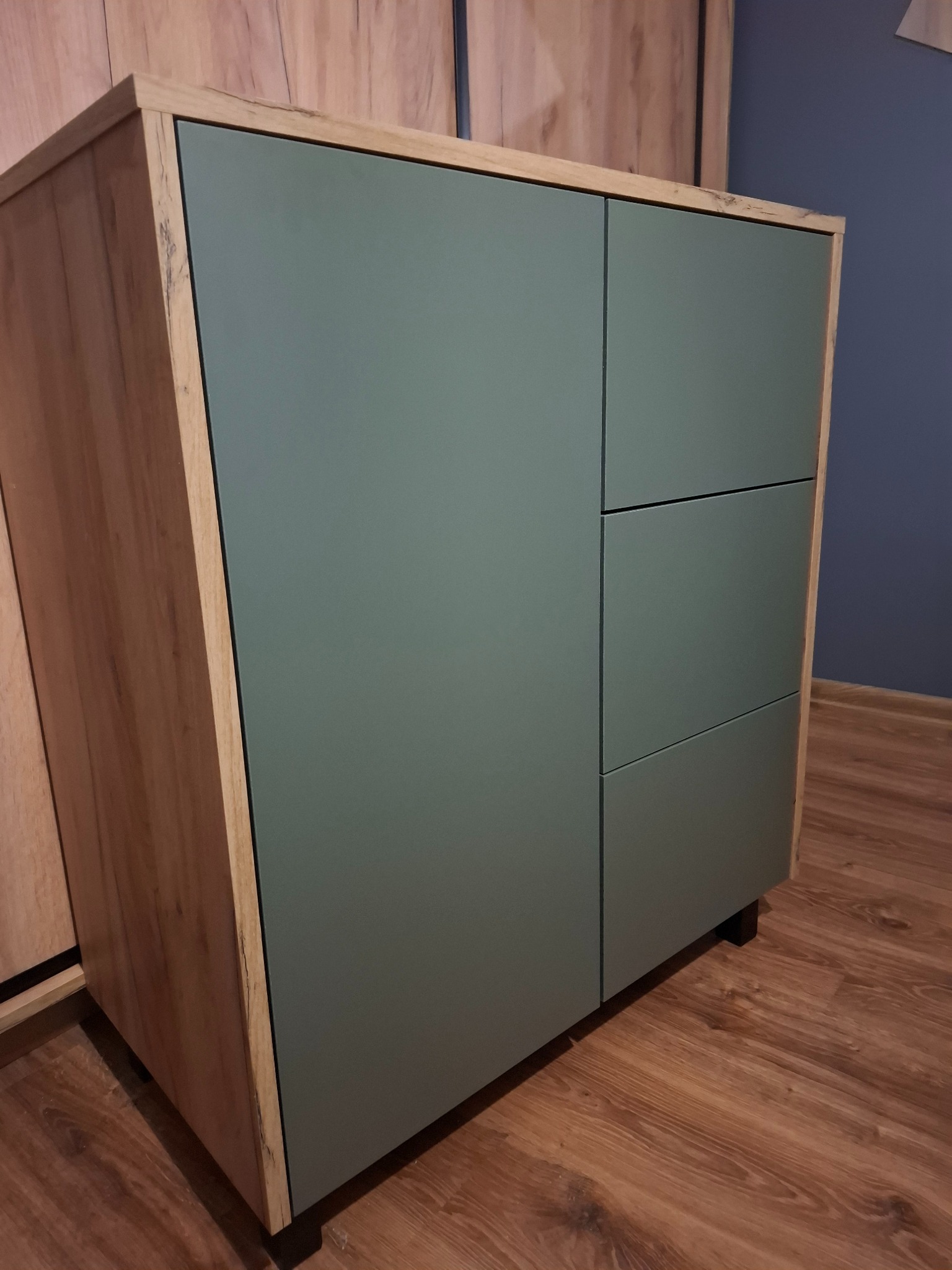 Niska szafka z dwiema pionowymi szafkami i trzema szufladami w kolorze zielonym, z obudową z drewna w stylu rustykalnym, na tle drewnianej podłogi i ściany w odcieniach niebieskiego.