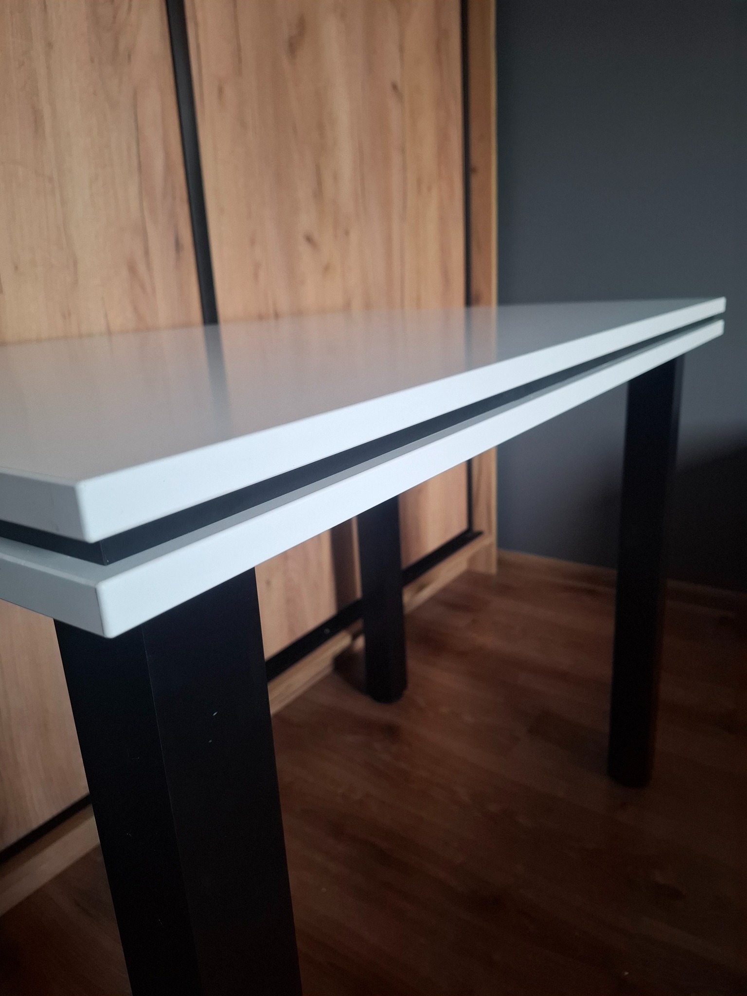 Biały stół z czarnymi nogami na tle szafy z drewnianymi frontami i szarej ściany, widoczne detale konstrukcyjne blatu.