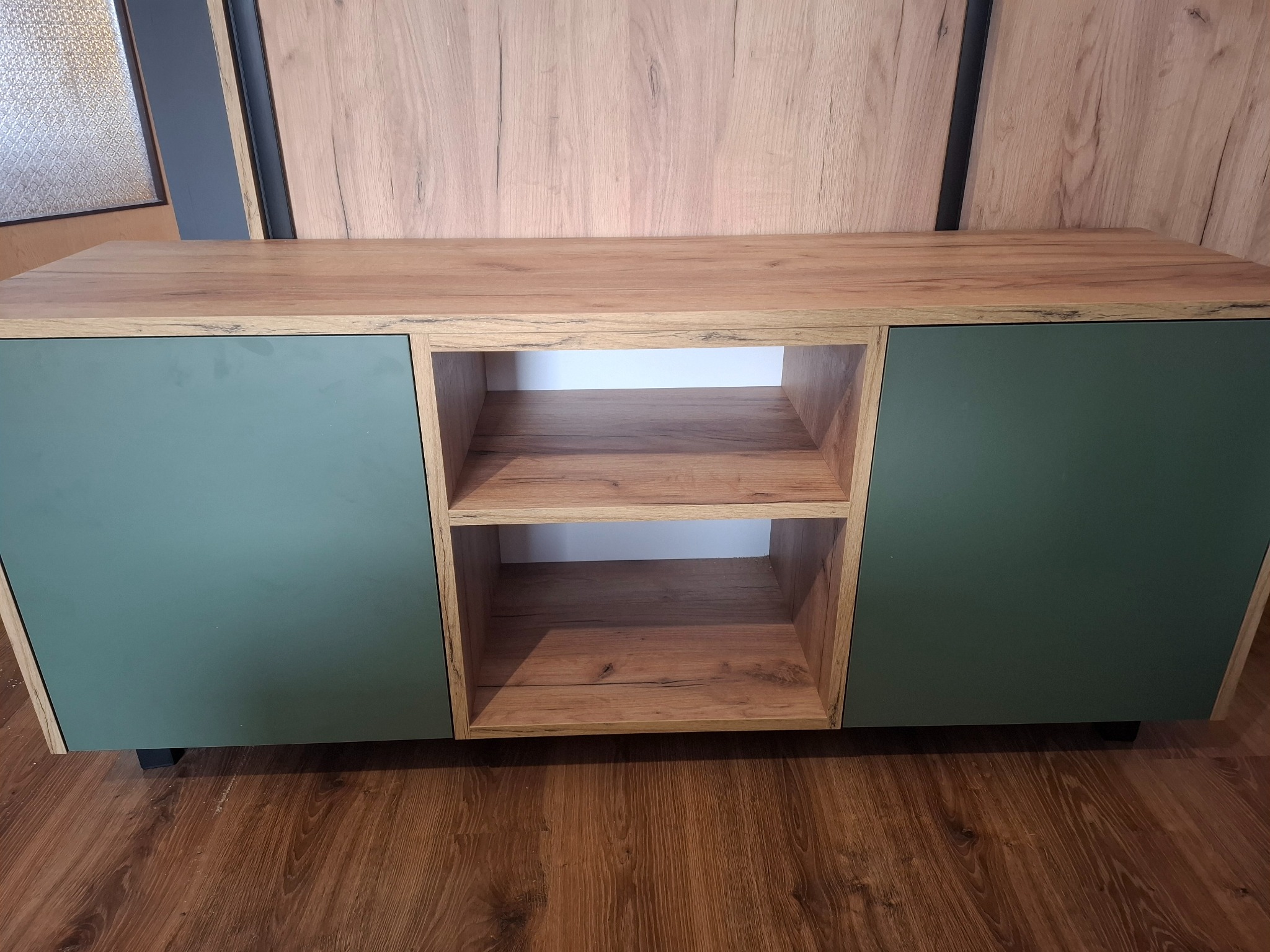 Nowoczesna szafka RTV z drewnianym blatem i zielonymi frontami, widoczne dwie otwarte półki i brązowa podłoga.