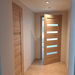 Banasiak kompleksowe wykończenia wnętrz - Korytarz z dwiema zamontowanymi drzwiami wewnętrznymi z poziomymi przeszkleniami z mlecznego szkła, oświetlony trzema punktowymi lampami sufitowymi, z podłogą wyłożoną panelami imitującymi drewno.