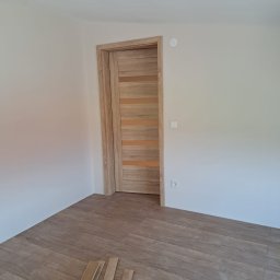 Banasiak kompleksowe wykończenia wnętrz - Jasne, drewnopodobne panele podłogowe i drzwi wewnętrzne z poziomymi przeszkleniami w nowym pomieszczeniu, widoczne listwy przypodłogowe i puszka elektryczna.