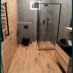 Banasiak kompleksowe wykończenia wnętrz - Nowoczesna łazienka z kabiną prysznicową typu walk-in z czarną ramą, umywalką nablatową w kształcie misy, drewnopodobną podłogą i szarymi płytkami ściennymi.