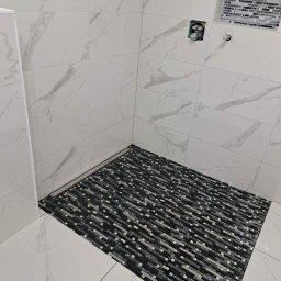 Banasiak kompleksowe wykończenia wnętrz - Ukończona kabina prysznicowa z białymi płytkami imitującymi marmur na ścianach i mozaiką w odcieniach szarości na podłodze oraz w półce ściennej, widoczny odpływ liniowy.