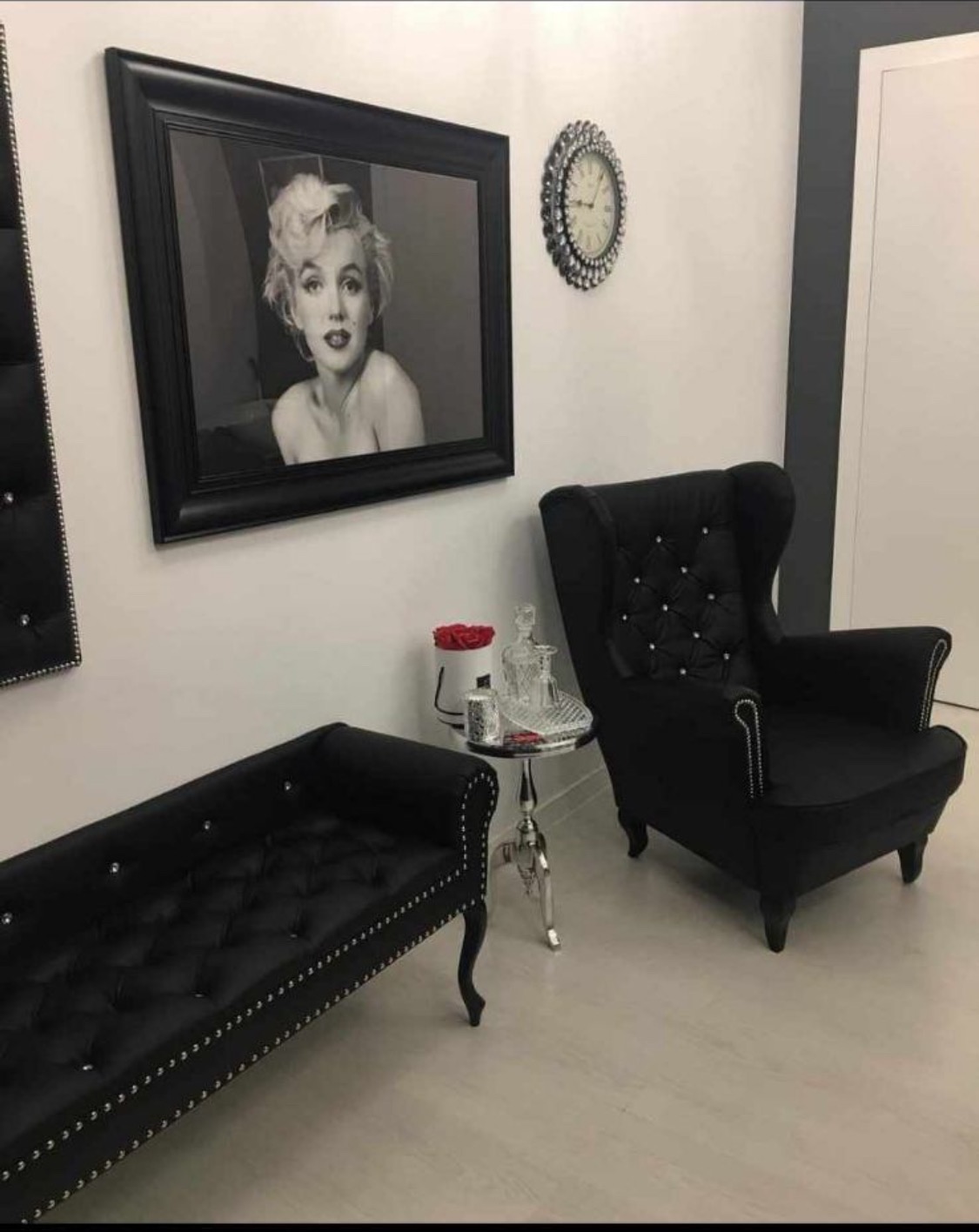 Elegancki czarno-biały wystrój wnętrza z czarnym pikowanym siedziskiem, fotelem typu uszak, srebrnym stolikiem, zegarem z kryształkami i portretem Marilyn Monroe w czarnej ramie.