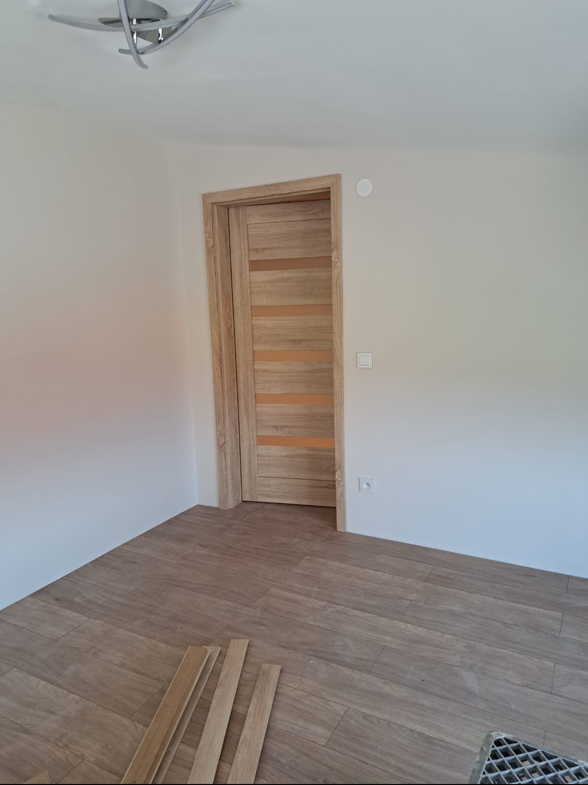Jasne, drewnopodobne panele podłogowe i drzwi wewnętrzne z poziomymi przeszkleniami w nowym pomieszczeniu, widoczne listwy przypodłogowe i puszka elektryczna.
