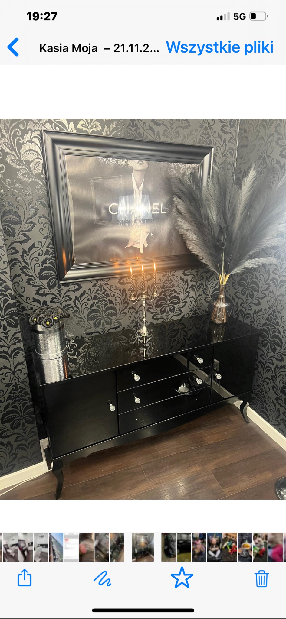 Elegancka aranżacja wnętrza w Łęczycy z czarną komodą, ozdobnym plakatem Chanel w ramie, świecznikiem i wazonem z pampasową trawą na tle czarno-srebrnej tapety.