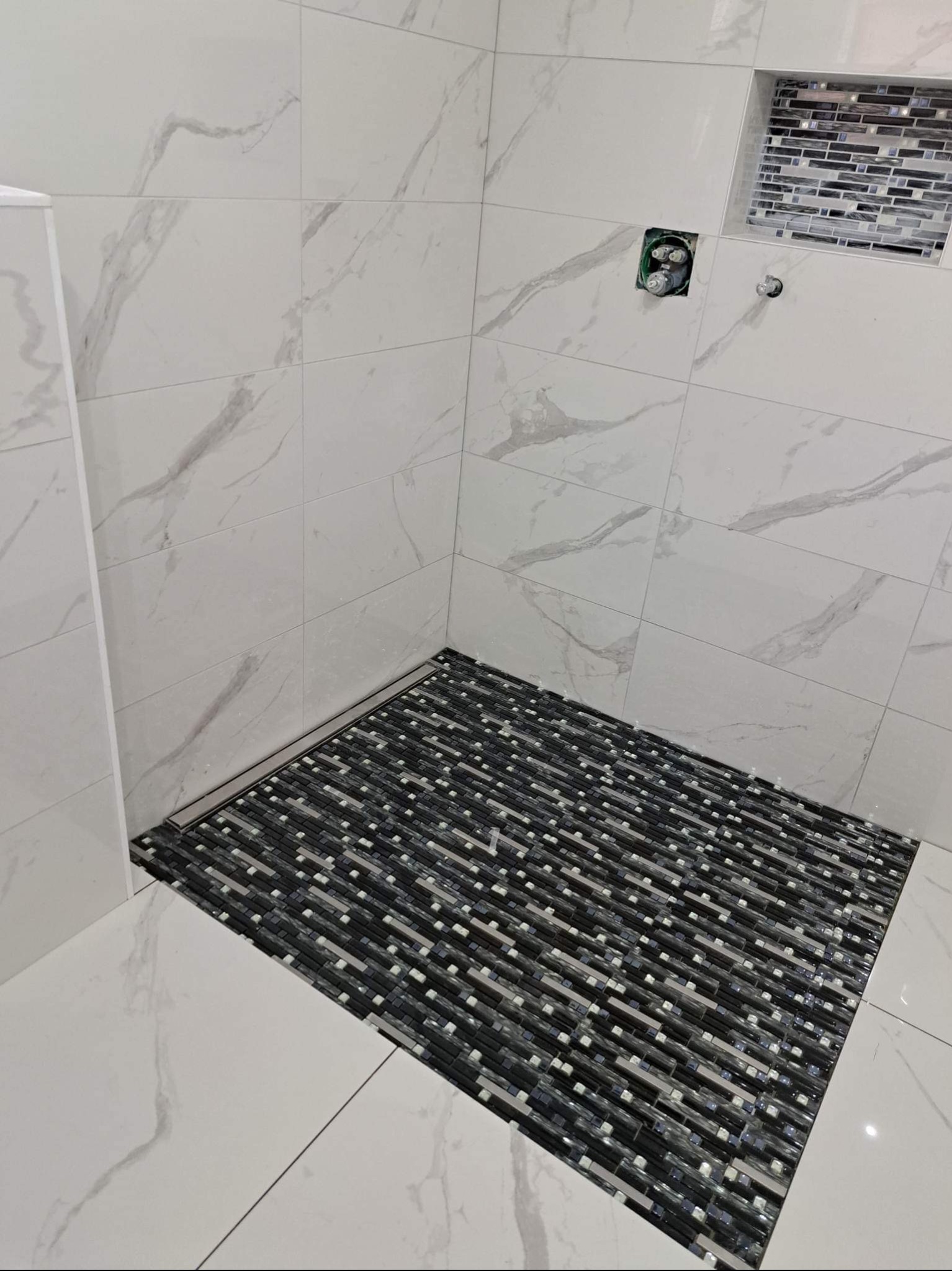 Ukończona kabina prysznicowa z białymi płytkami imitującymi marmur na ścianach i mozaiką w odcieniach szarości na podłodze oraz w półce ściennej, widoczny odpływ liniowy.