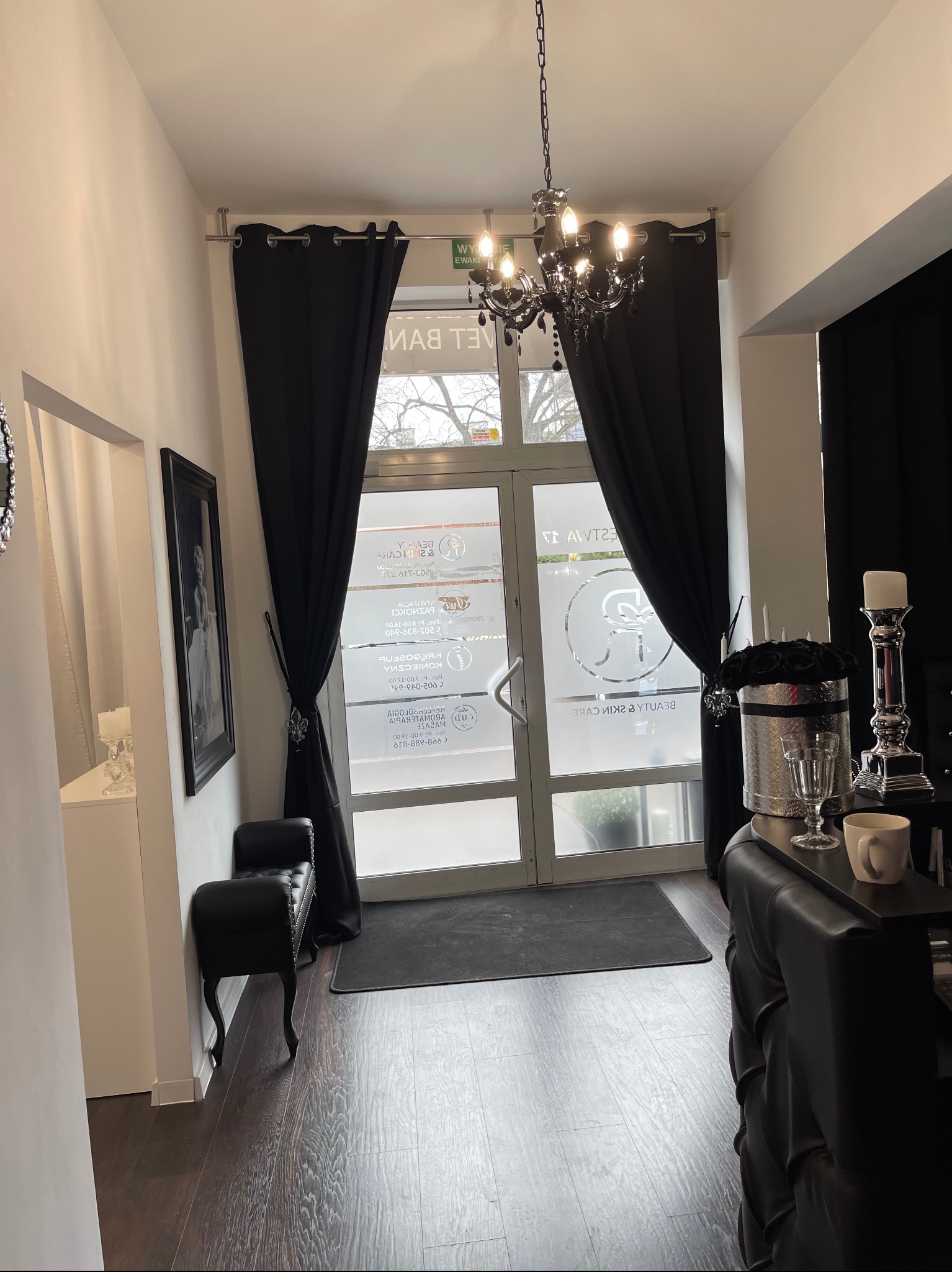 Eleganckie wejście do salonu kosmetycznego z ciemnymi zasłonami, czarnym żyrandolem i dekoracjami w srebrnych odcieniach, widoczne logo firmy na szklanych drzwiach.