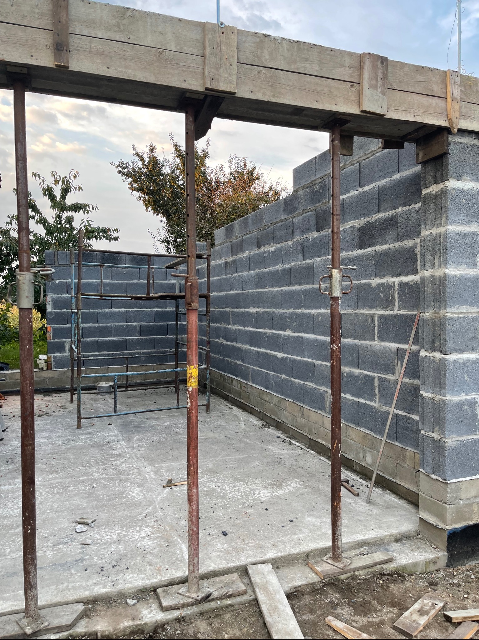 Wznoszenie ścian z szarych bloczków betonowych, podparte drewnianą konstrukcją i metalowymi stemplami, widoczny fragment rusztowania i betonowa podstawa.