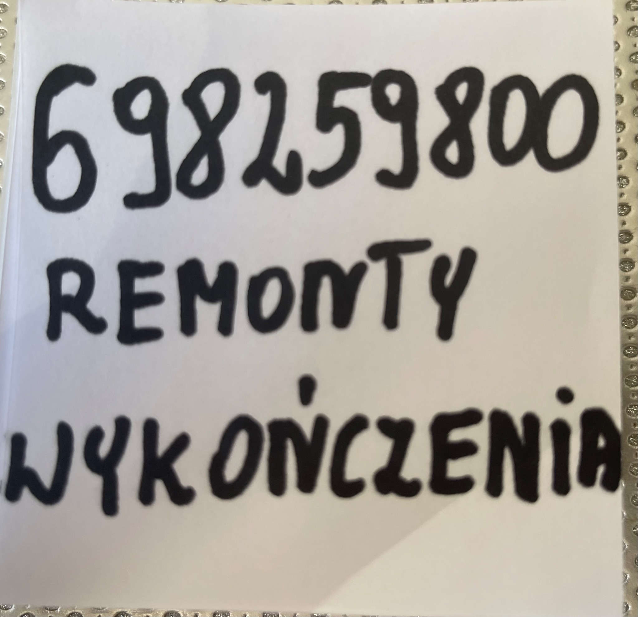 Kartka z numerem telefonu 698259800 i napisem 'Remonty Wykończenia' napisanym markerem, leżąca na srebrnej, błyszczącej powierzchni.