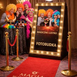 Magia w Obiektywie - Uśmiechnięci ludzie w perukach pozują w fotobudce z napisem 'Magiczne Lustro Fotobudka', na czerwonym dywanie z logo firmy. Zabawna atmosfera imprezy.